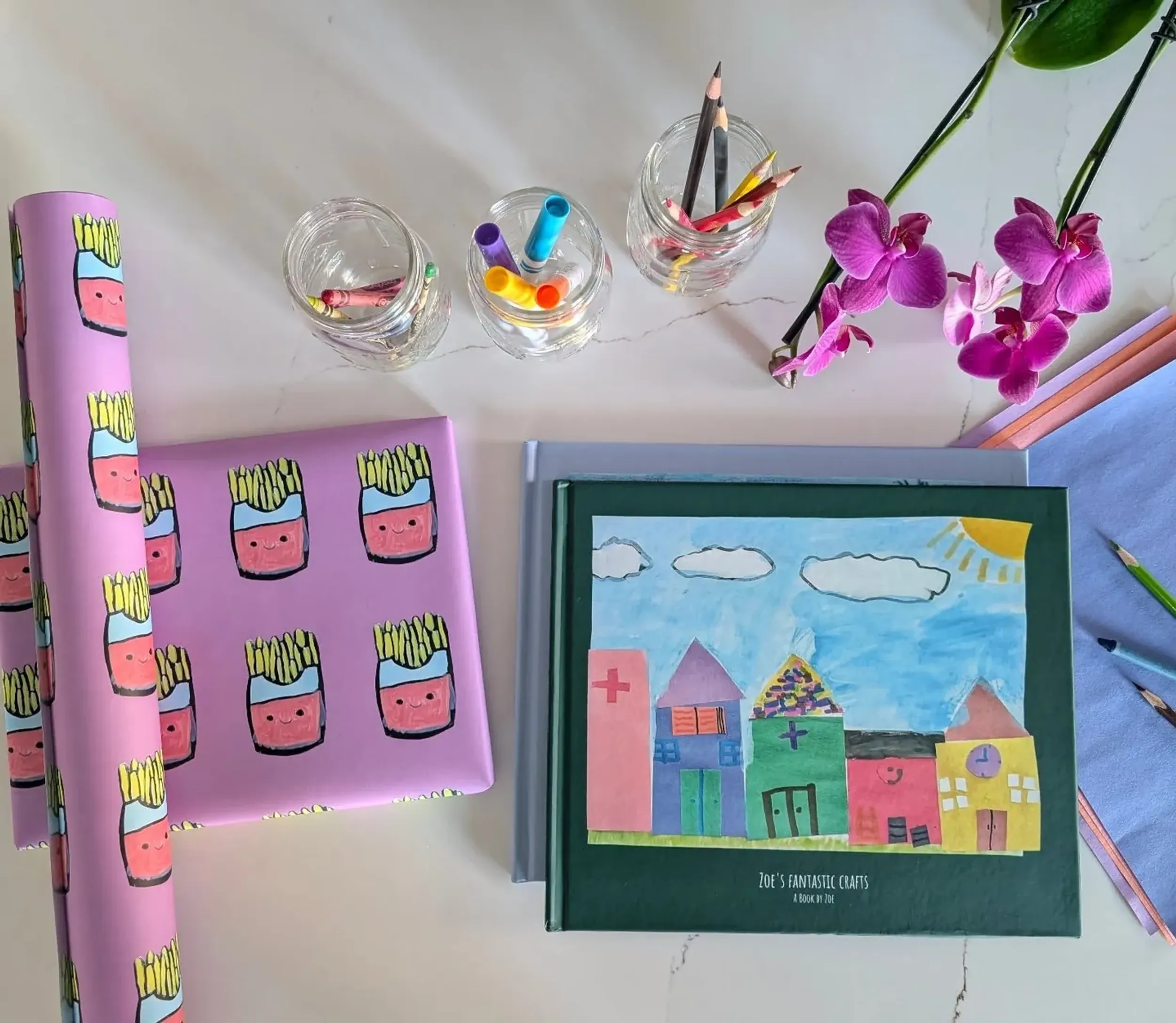 Kids art gift wrap printed with a child's cartoon french fry characters, shown with a hardcover kids art book and colored pencils