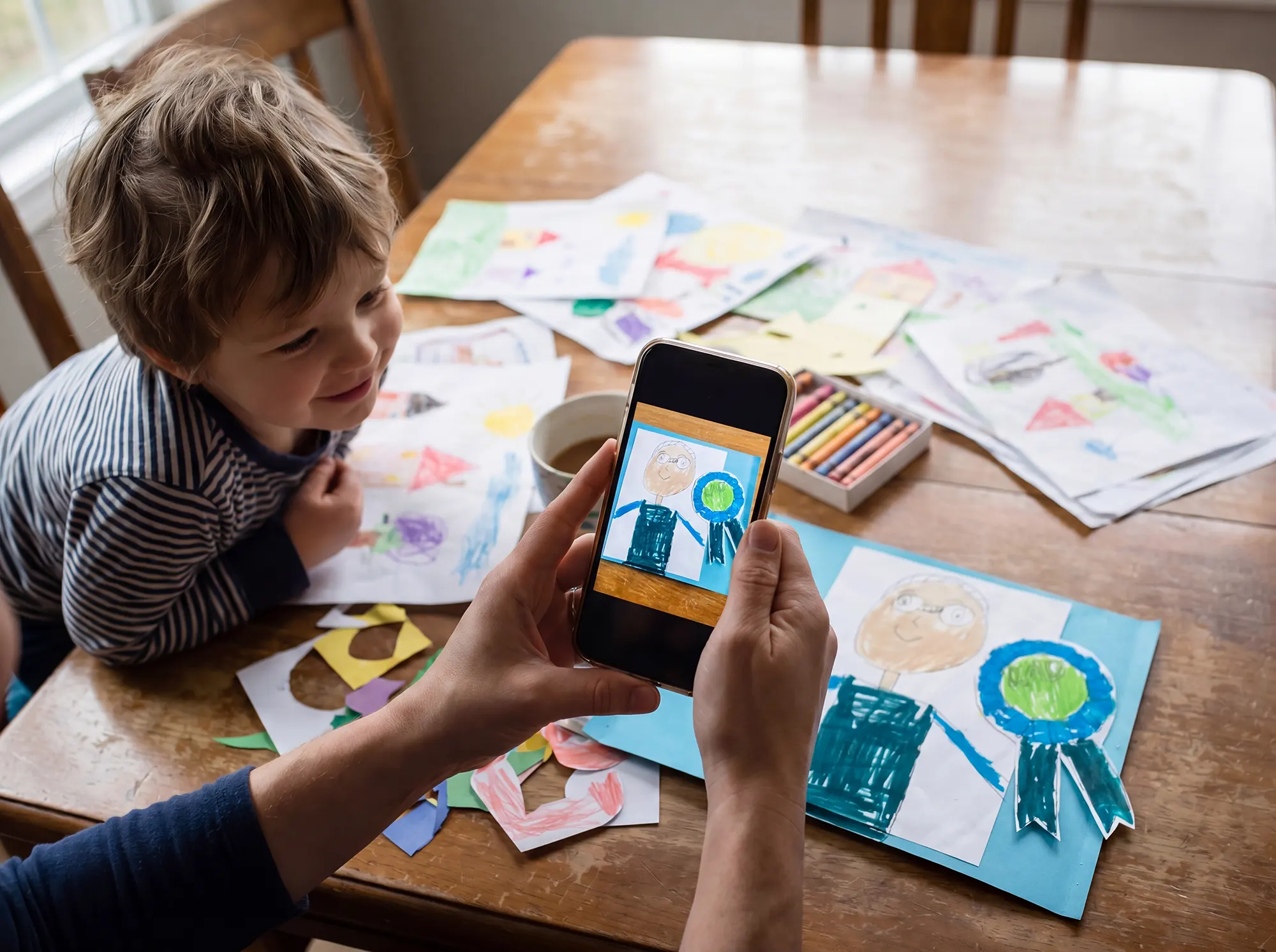 A mom photographs her child's artwork with her phone at a kitchen table covered in colorful kids artwork, as her young son watches.