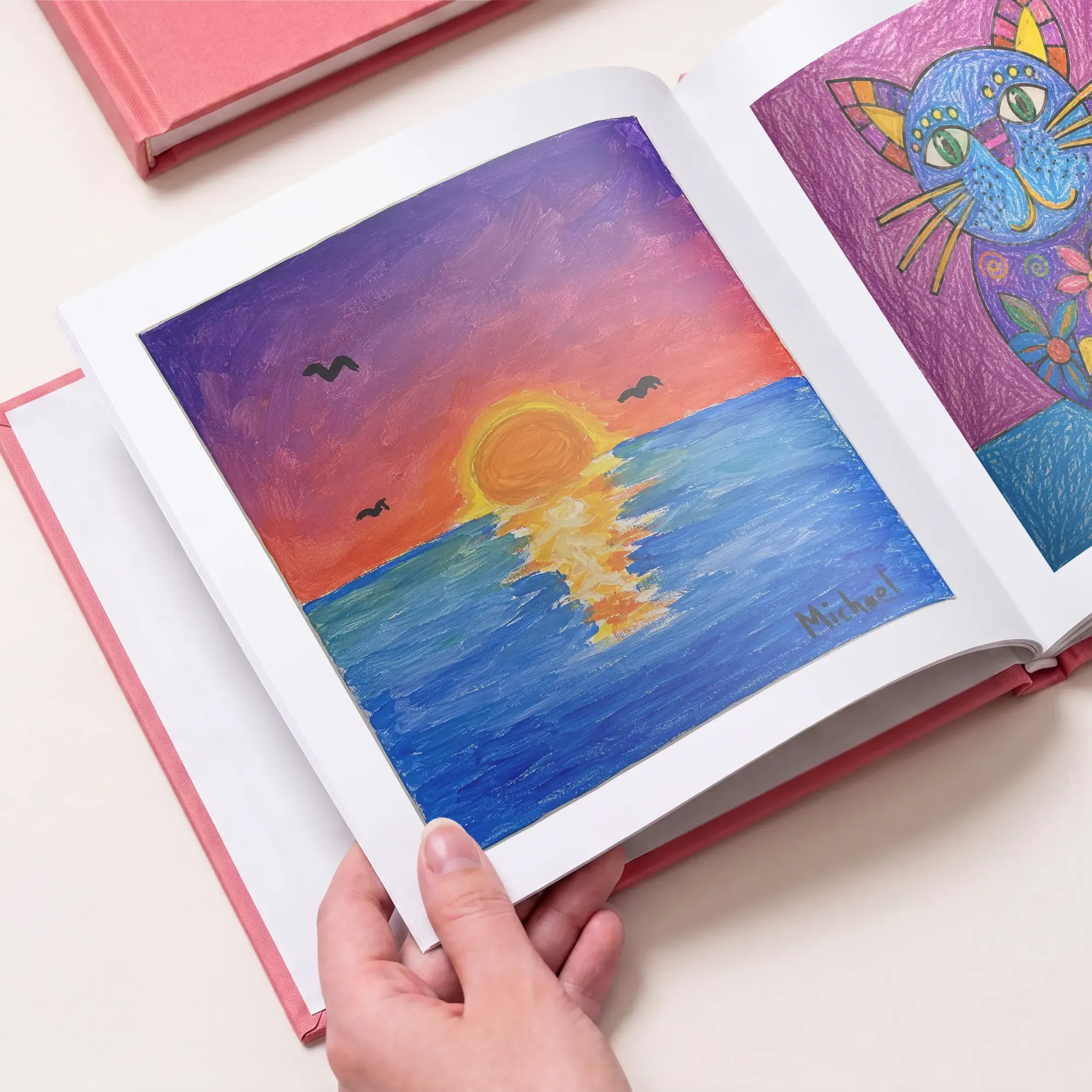 Inside a kids art book showing a child's painted sunset over the ocean and a colorful cat illustration, one artwork per page